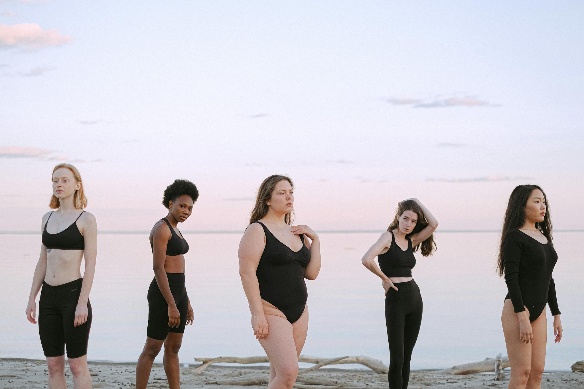 Five women of diverse backgrounds posing confidently on a serene beach.